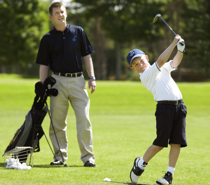 a kid swinging his golf club with his dad looking at him from behind