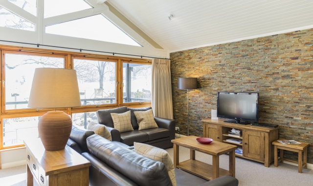 Cameron House bungalow living room, with leather sofa, brown coffee table and TV unit. Loch Lomond is visible through the windows.