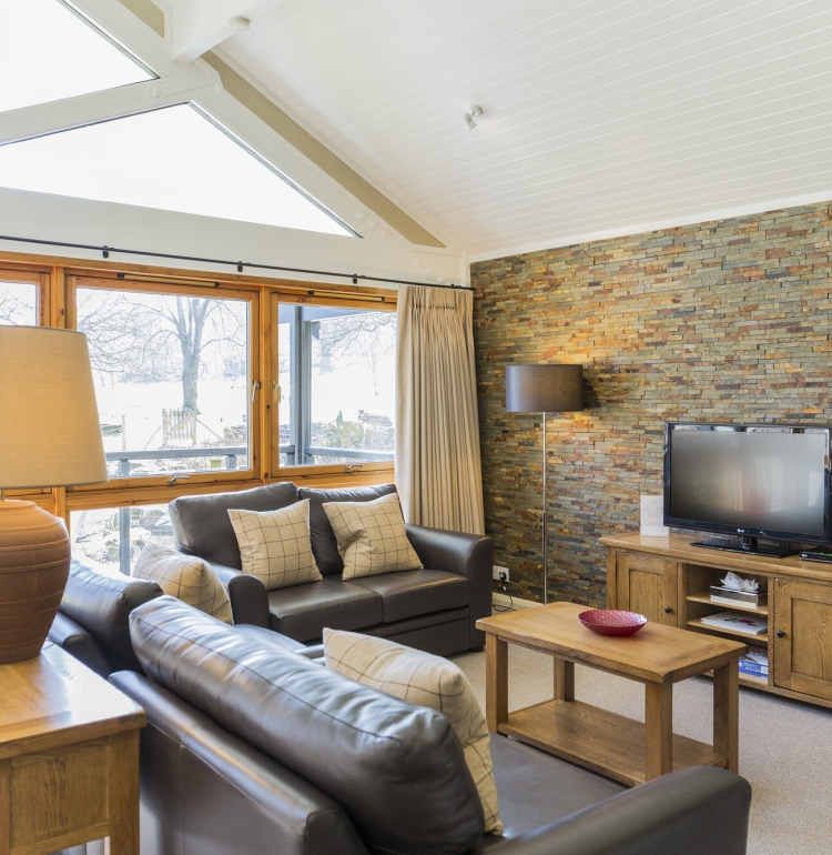 Cameron House bungalow living room, with leather sofa, brown coffee table and TV unit. Loch Lomond is visible through the windows.