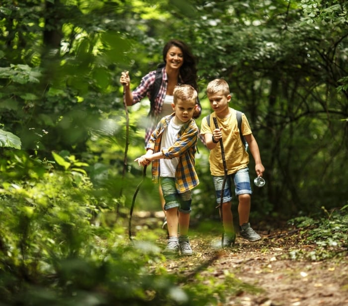 a woman walking with two small kids using sticks to walk down a dirt trail in the woods