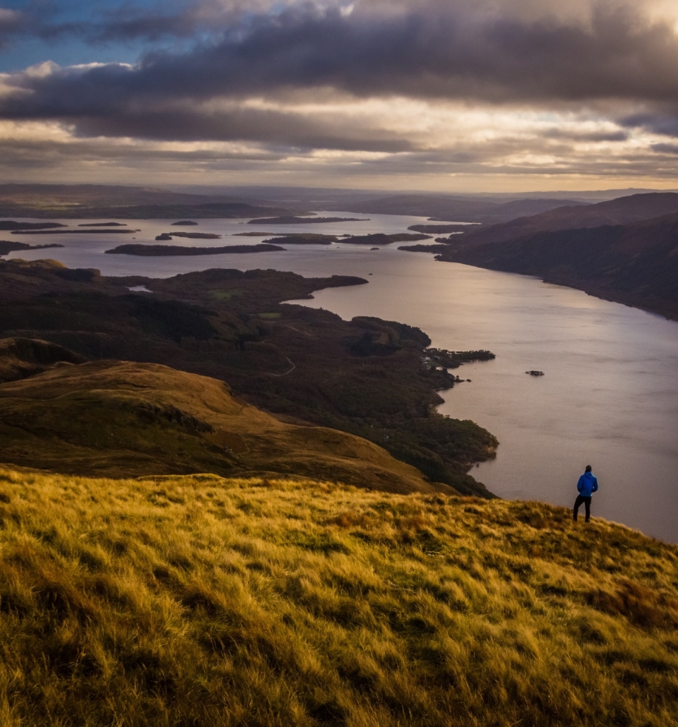 a person standing on the side of a hill over looking a body of water and other hills