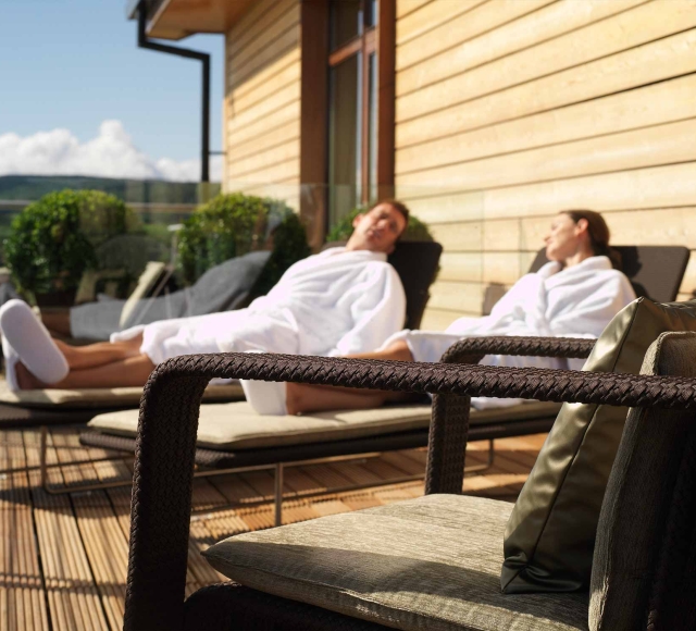 a male and female sitting outside in robes on lounge chairs enjoying the sun