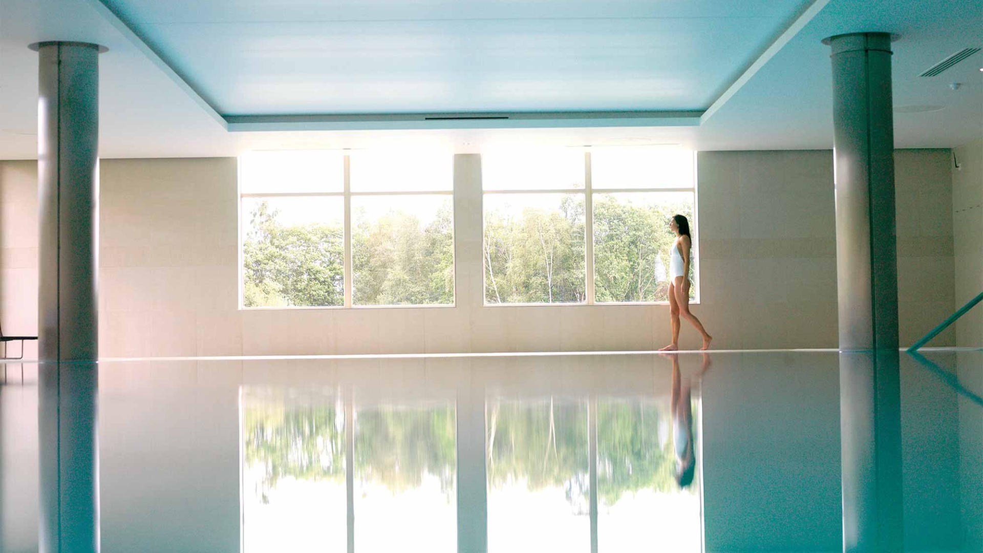 a woman walking along the side of a indoor pool looking outside the window