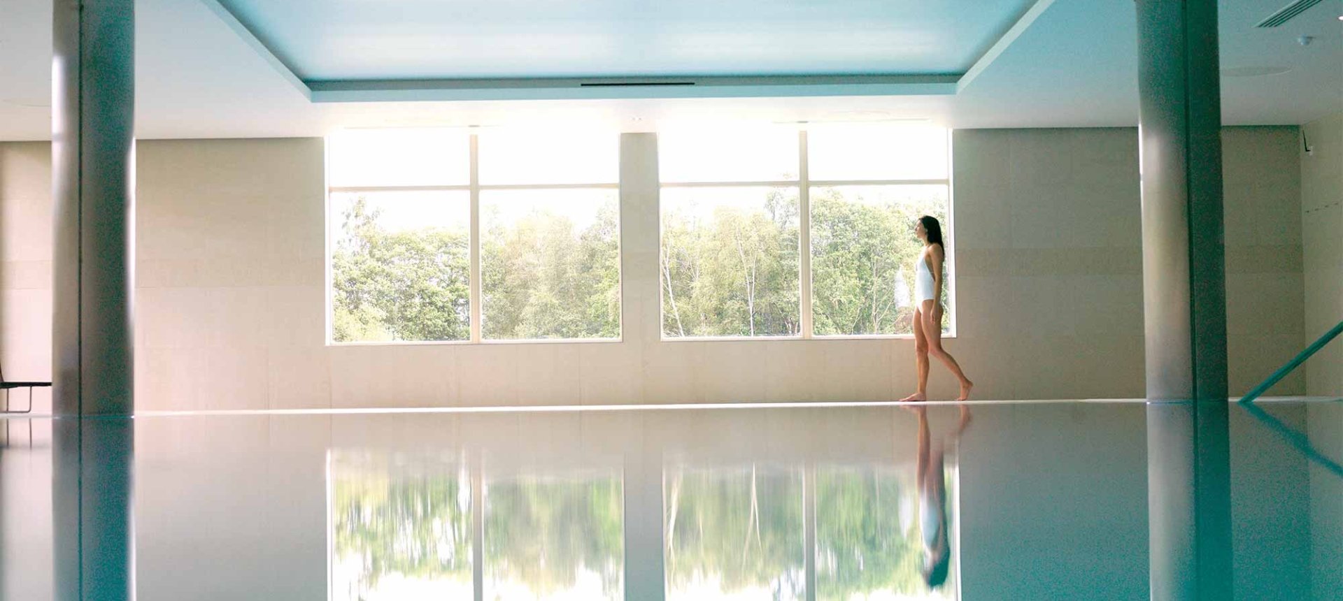 a woman walking along the side of a indoor pool looking outside the window