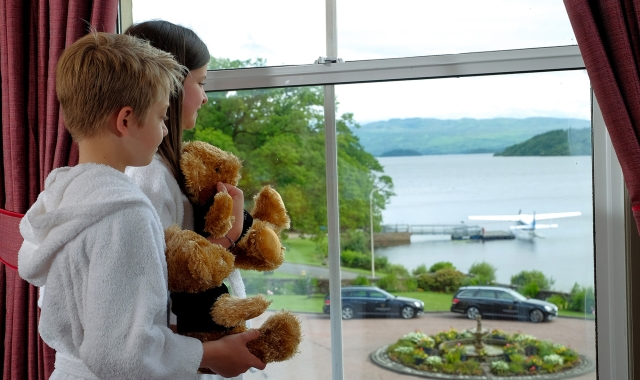 two kids holding teddy bears looking out a window at the water