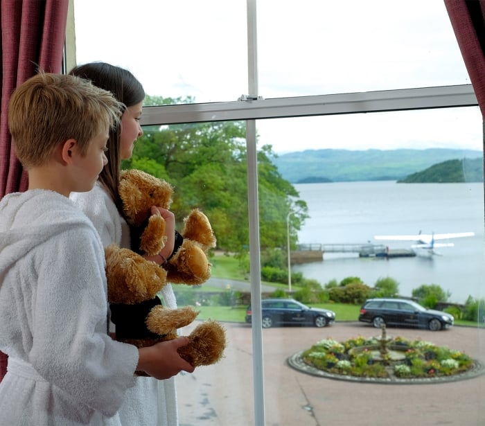 two kids holding teddy bears looking out a window at the water