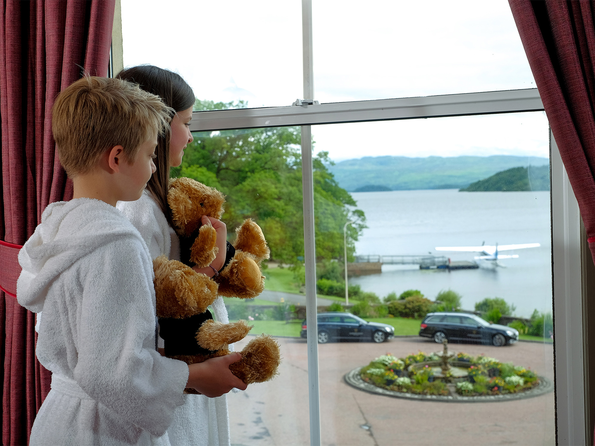 two kids holding teddy bears looking out a window at the water