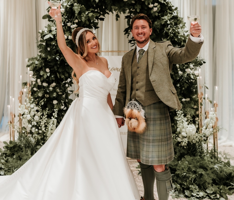 A bride and groom standing at a floral archway holding up their hands in celebration