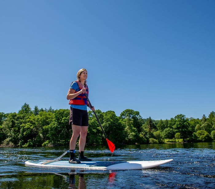 a woman standing outside on a surfboard holding red and black paddle in the middle of a lake