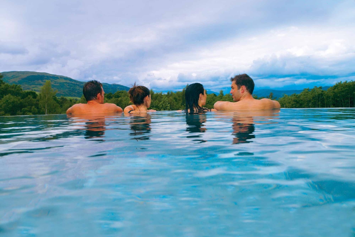 two couples sitting in an out door infinity pool looking over the edge to the forest below