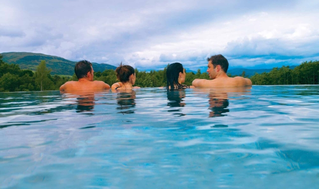 two couples sitting in an out door infinity pool looking over the edge to the forest below