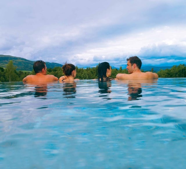 two couples sitting in an out door infinity pool looking over the edge to the forest below