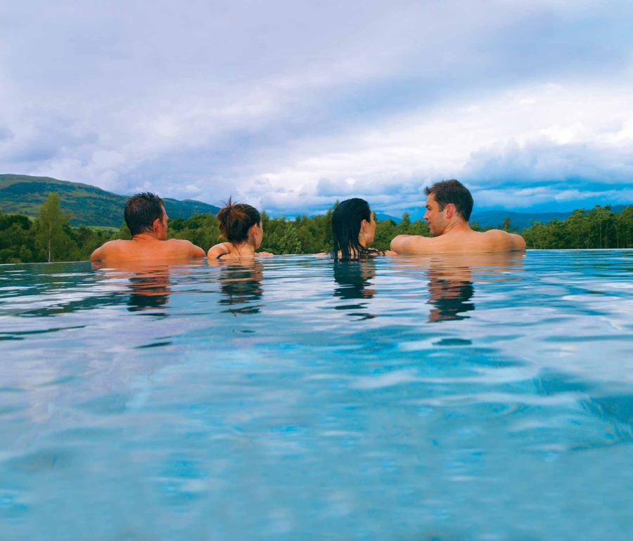 two couples sitting in an out door infinity pool looking over the edge to the forest below