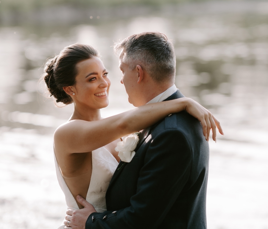 A bridge and groom standing outside in front of a lake in their wedding outfits