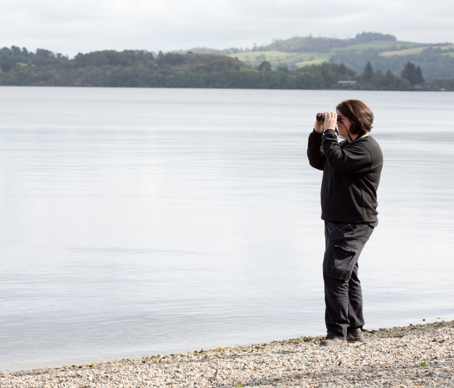 a women dressed all in black stands at the end of Loch Lomond, looking through binoculars.