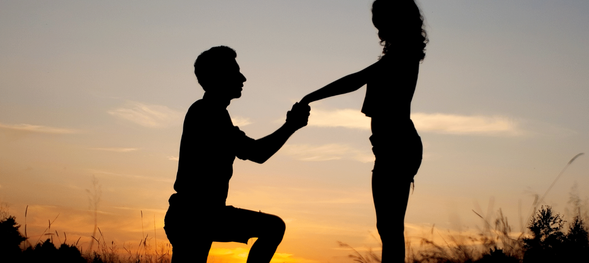 a man kneeling down holding a woman's hand while the sun sets behind them