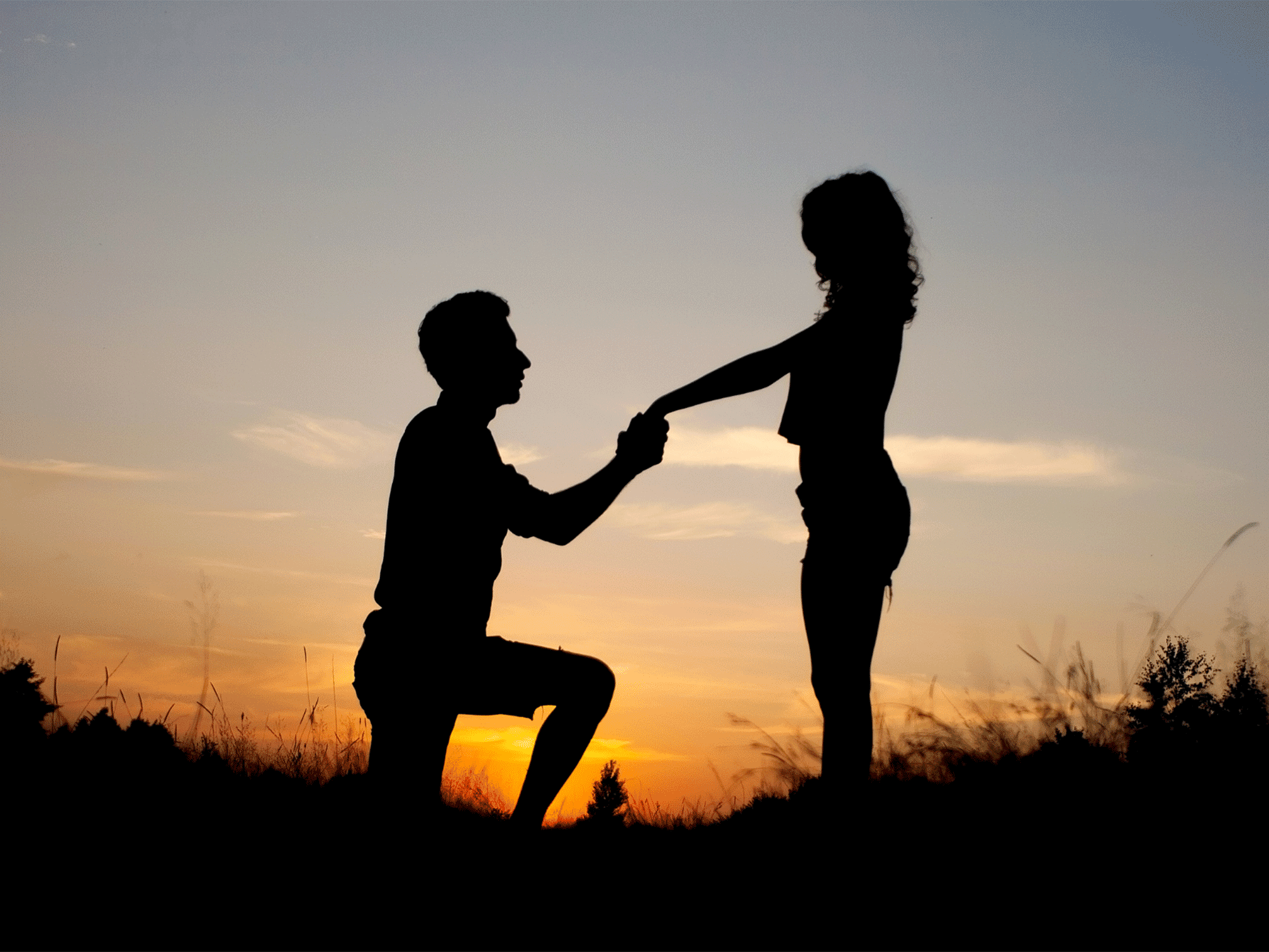 a man kneeling down holding a woman's hand while the sun sets behind them