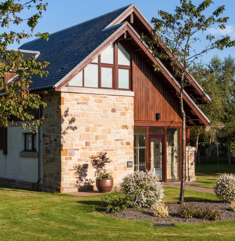 A modern, luxury detached self-catering Loch Lomond lodge with Scottish architectural features. The two-story exterior combines warm wood paneling and rustic golden stone cladding under a steep slate roof. The lodge is set within a well-manicured green lawn and surrounded by mature trees, suggesting a tranquil holiday setting on the shores of Loch Lomond.