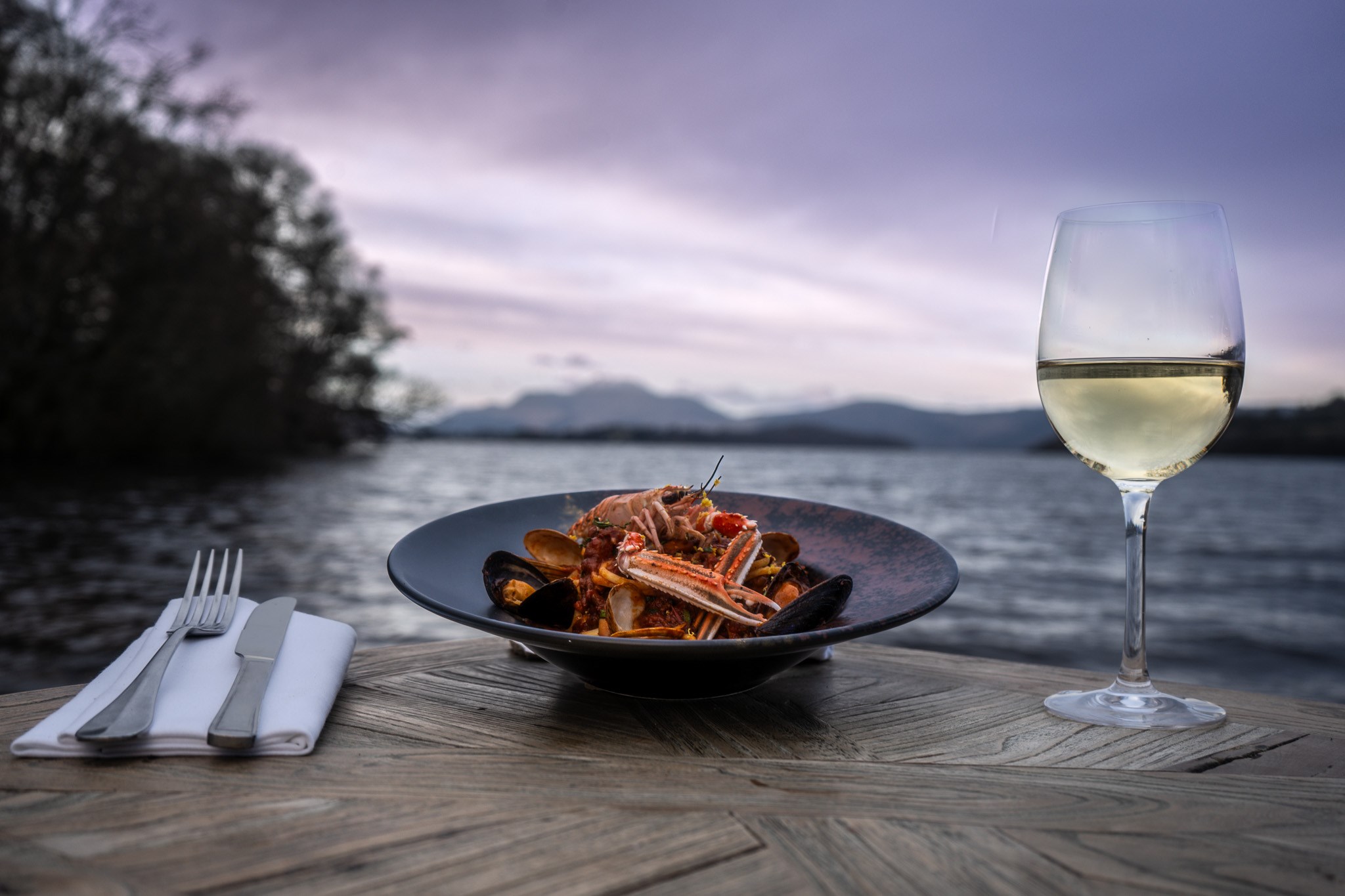 a beautiful seafood dish sitting next to silverware and champagne out doors in front of the water