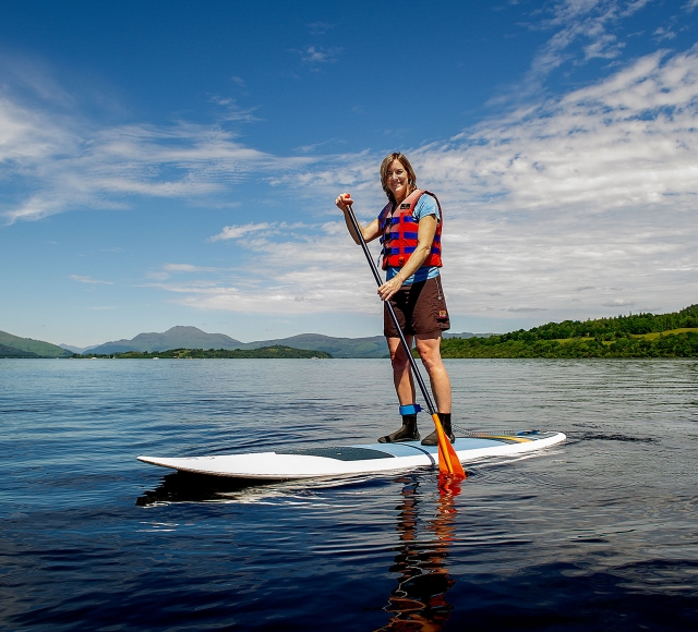 a woman standing on a paddle board in the middle of the water with bright green trees and hills behind her