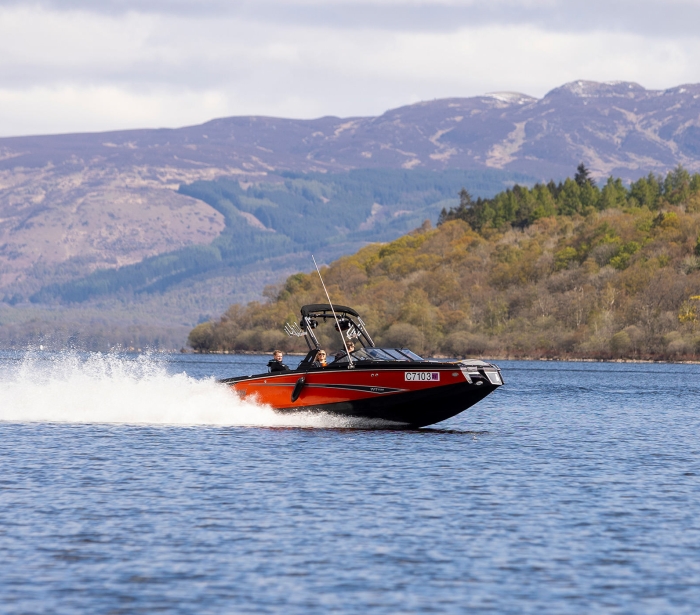 people riding on a red and black boat in the middle of the water with mountains in the background