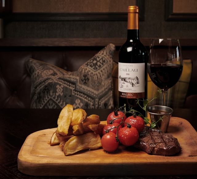 a board with steak, tomatoes, potatoes and red wine