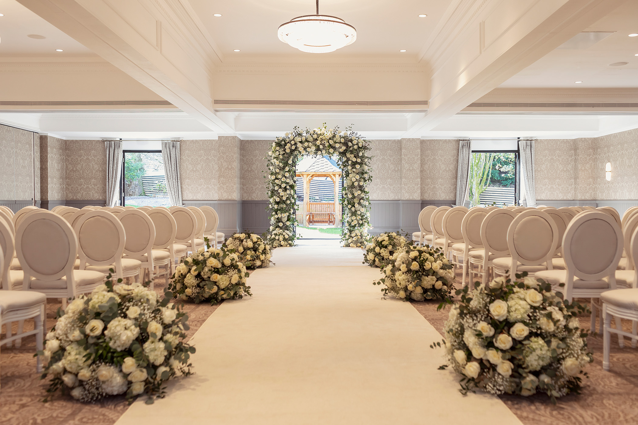 a walking space with a white carpet and flower bushes on each side leading to a outdoor gazebo