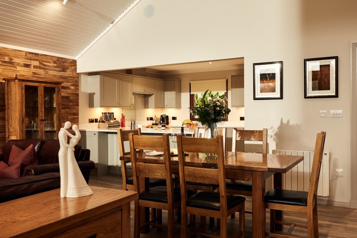 Dining area with a wooden table and. chairs. in the background is the kitchen area where meals can be prepared.