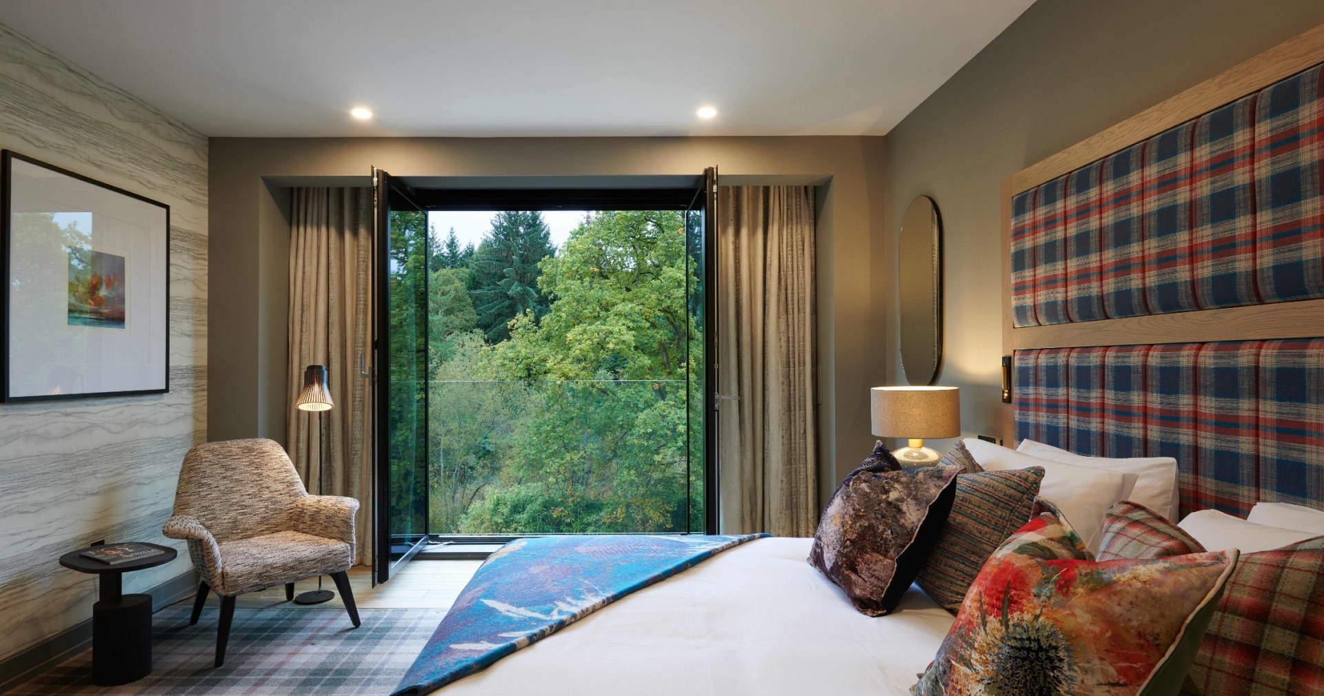 Hotel room with bed, chair, and view of lush green trees from window.