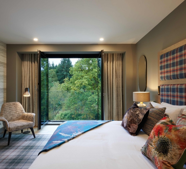 Hotel room with bed, chair, and view of lush green trees from window.