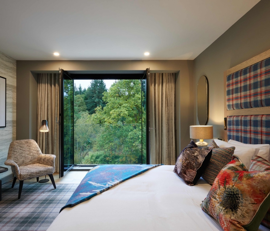 Hotel room with bed, chair, and view of lush green trees from window.