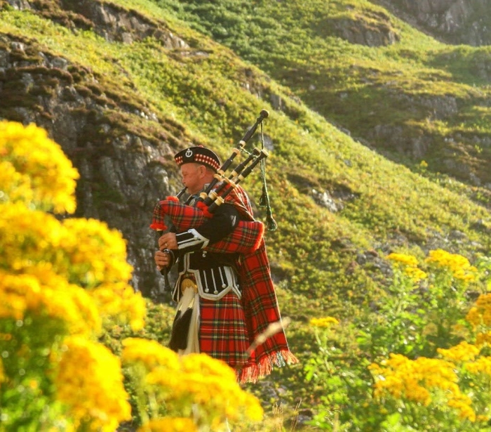 A Scottish piper dressed in kilt, piping in the Scottish hills during a sunny day.